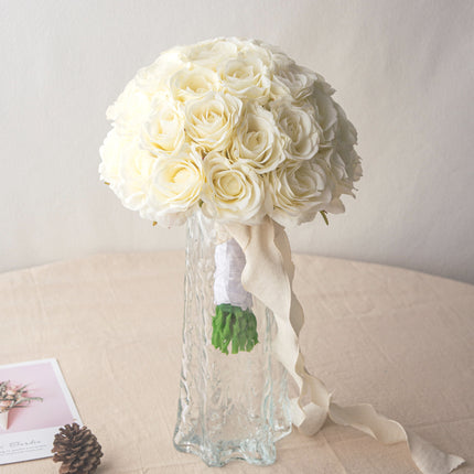 High-Quality artificial cream white silk rose bridal bouquet in a glass vase with ribbon on a table.