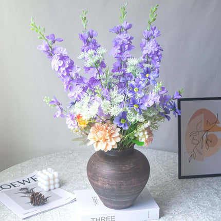 Handmade delphinium artificial flower arrangement in a dark vase with assorted flowers on a table.